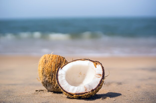 반으로 쪼개진 아름다운 익은 코코넛은 푸른 바다와 구름이 있는 푸른 하늘의 흐릿한 배경에 있는 하얀 모래 해변에 놓여 있습니다