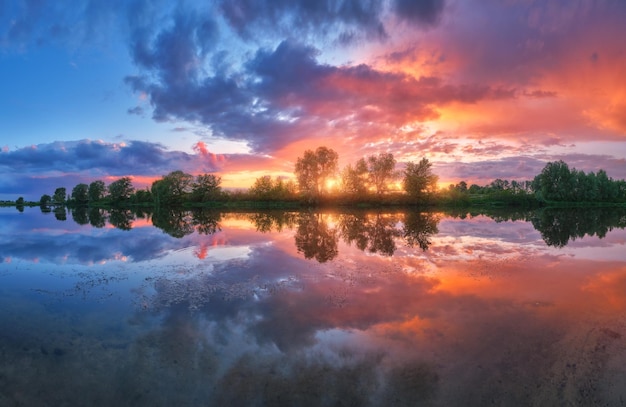 Beautiful river coast at sunset in summer Colorful landscape with lake green trees and grass blue sky with multicolored clouds and orange sunlight reflected in water Nature Vibrant scenery