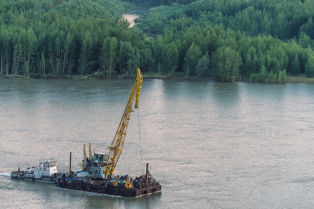 Beautiful river landscape with green shores and ship with copy space. Towboat tows barge with crane along riverbank. Dawn reflexed on calm glare water surface.
