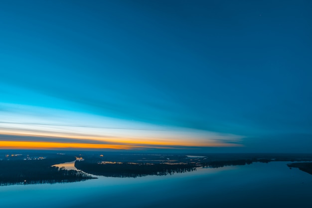 Beautiful river with big island with trees under predawn sky. Bright orange stripe in picturesque cloudy sky. Early blue sky reflected in water. Colorful morning atmospheric image of majestic nature.