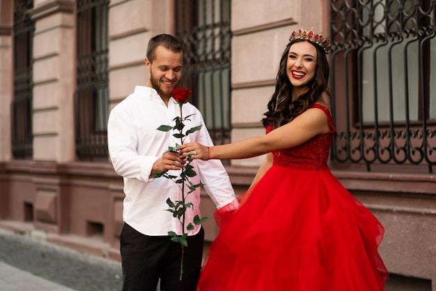 美しいロマンチックなカップル。赤いドレスと路上を歩く赤いバラと白いシャツにハンサムな男と王冠の魅力的な若い女性。幸せな聖バレンタインの日。妊娠中や結婚式のコンセプトです。