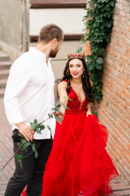 Beautiful romantic couple. Attractive young woman in red dress and crown with handsome man in white shirt with red rose walking on the street. Happy Saint Valentine's Day. Pregnant and wedding concept.