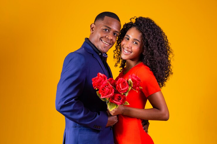Beautiful romantic couple isolated on yellow background Attractive young woman in red dress holding red roses and handsome man in a suit are in love Happy Valentine's Day