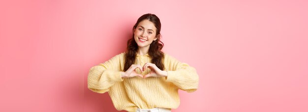 Photo beautiful romantic girl say i love you showing heart sign and smiling at camera standing cute agains