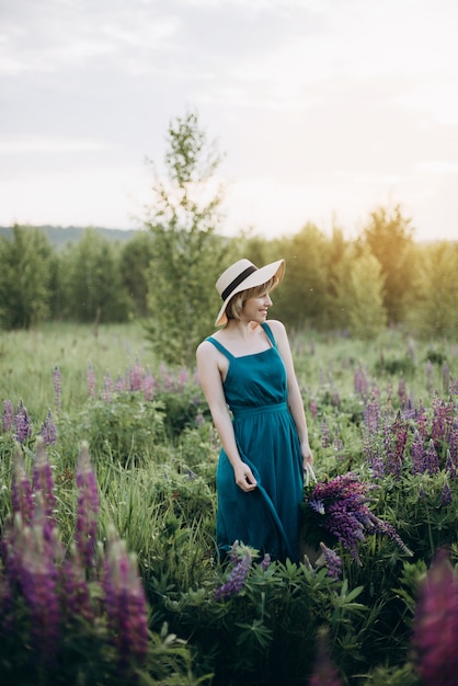 Beautiful romantic woman blonde in a dress and hat in a field with a bouquet of purple flowers of lupins at dawn .