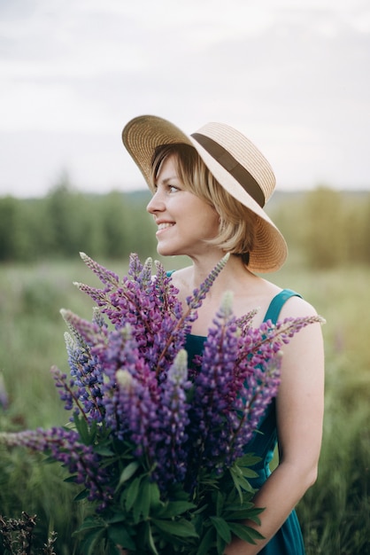 A beautiful romantic woman smiles in a dress and hat in a flower field with a huge bouquet of lupine flowers.