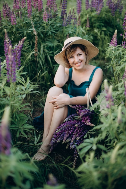 A beautiful romantic woman smiles joyfully in a blue dress and hat sits in a field of purple lupine flowers.