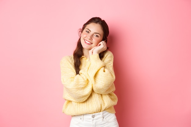 Beautiful romantic woman with bright make up and spring outfit looking happy and delighted at camera holding hands near face standing over pink wall