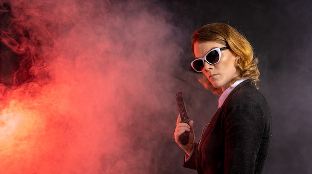 beautiful rough elegant woman with gun in hand. Smoke and fire on dark background.