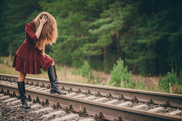 Beautiful sad woman in crimson dress is hiding face by hair in forest on railway