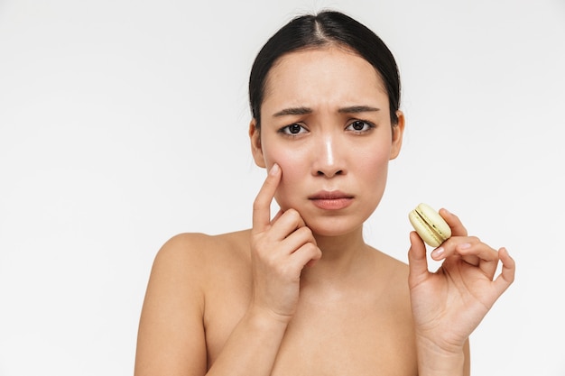 Photo beautiful sad young pretty asian woman with healthy skin posing naked isolated over white wall holding sweeties macaroons.