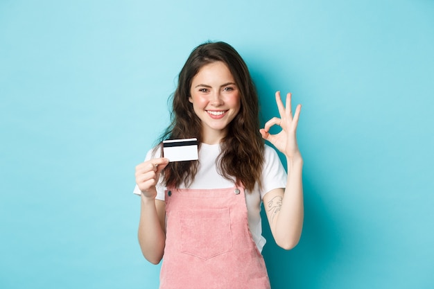 Beautiful sassy girl recommending plastic credit card, showing okay sign and looking confident at camera, smiling satisfied, standing over blue background