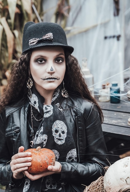 Красивая страшная маленькая девочка празднует хэллоуин Ужасающий черно-белый макияж на половину лица Костюм ведьмы стильный образ Ужасное веселье на детской вечеринке в сарае на улице Шапка куртка тыква