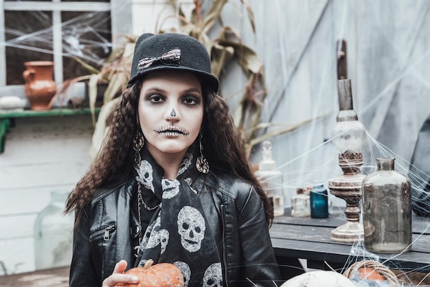 Beautiful scary little girl celebrating halloween Terrifying black white halfface makeupwitch costume stylish image Horror fun at children's party in barn on street Hat jacket pumpkin