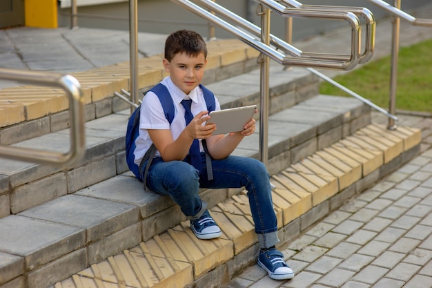 A beautiful schoolboy in a white shirt with a blue backpack, blue tie, blue jeans sits on the stairs outside and plays with a gray tablet.