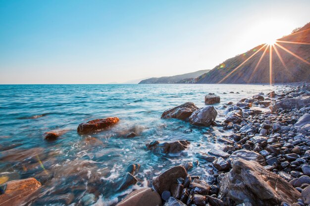 暖かい晴れた夏の朝、高い山や丘を背景に岩のビーチに沿って波がはねかける美しい海の景色。旅行のコンセプト。広告スペース