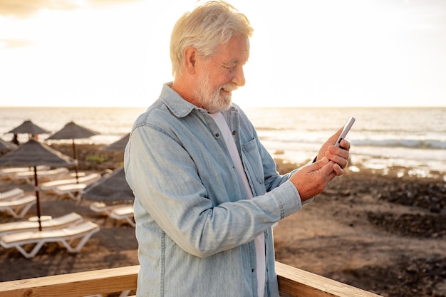 Beautiful senior bearded man in casual shirt standing outdoors at beach in sunset using phone