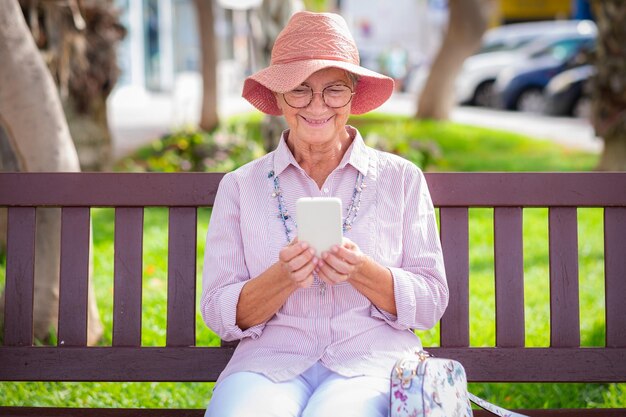 Beautiful senior caucasian woman wearing pink hat sitting on a bench using mobile phone happy retired people enjoying free time and technology