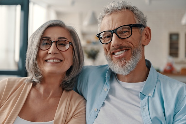 Beautiful senior couple in casual clothing smiling and looking at camera while spending time at home