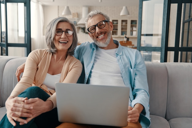 Beautiful senior couple in casual clothing smiling and using laptop while bonding together at home
