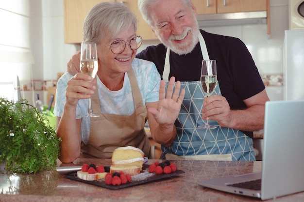 Beautiful senior couple in the home kitchen after baking their homemade plumcake they hold two glasses of wine while chatting with friends or family on the computer Cooking at home together