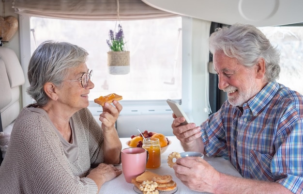 Beautiful senior couple in travel vacation leisure sitting\
inside a camper van enjoying breakfast together caucasian elderly\
husband using phone while the wife looks at him