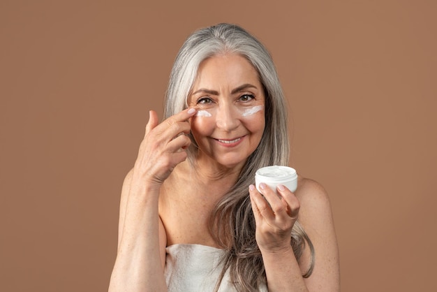 Beautiful senior woman applying moisturizing or nourishing cream under her eyes on brown studio