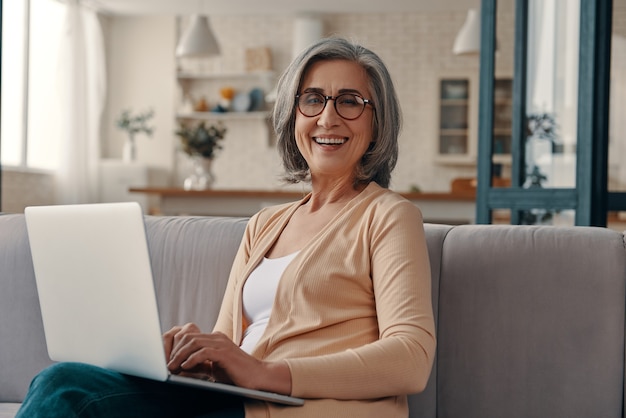 Beautiful senior woman in casual clothing using laptop and looking at camera while sitting on the sofa at home