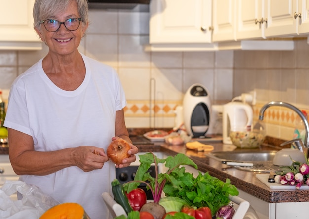 Beautiful senior woman in the home kitchen preparing vegetables. Fresh raw harvested on the table