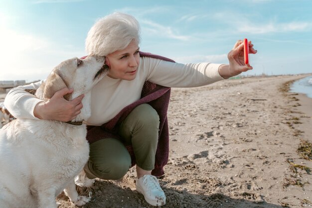 Beautiful senior woman photographing herself and the pet with the smartphone on the sandy beach