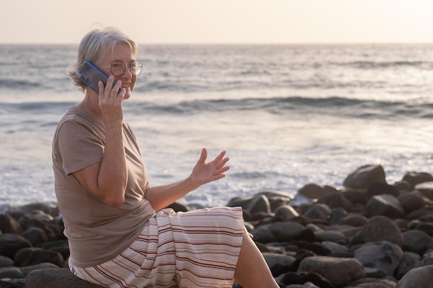 Bella donna anziana seduta in spiaggia al tramonto utilizzando smart phone sorridente anziana signora rilassata gode di vacanza e libertà