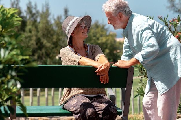 Beautiful senior woman sitting outdoor on a bench of public park talking with her husband Smiling elderly grandparents enjoy relax and retirement in sunny day