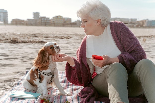 Beautiful senior woman with the smartphone sitting on the sand and stroking her cute puppy