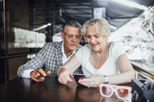 Beautiful seniors couple searching something at phone sitting in the summer terrace in modern cafe