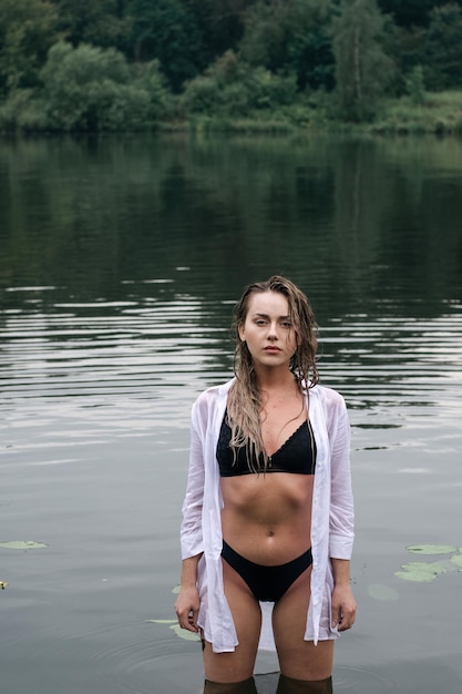 Beautiful sensual blonde girl with wet hair in a white shirt and black swimsuit posing in the lake.