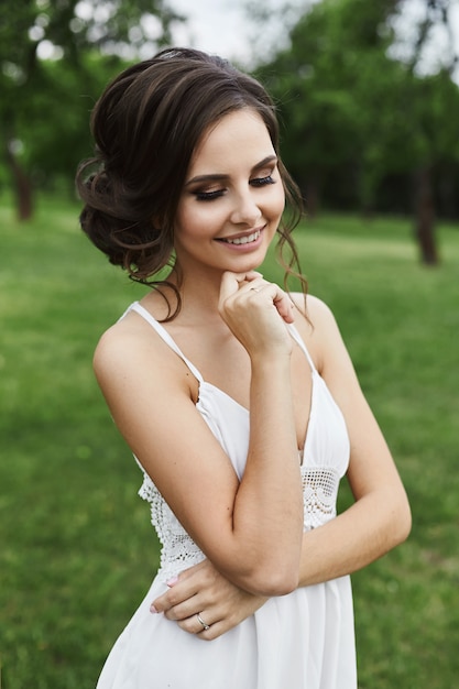 Bella e sensuale ragazza modello bruna in breve abito bianco, con gli occhi chiusi, con acconciatura alla moda e in posa al parco verde in prima serata