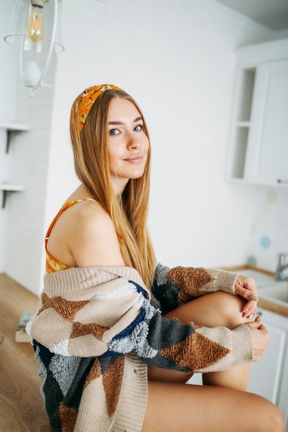 Beautiful sensual smiling young woman fair long hair girl wearing in the cozy knitted cardigan sitting on kitchen table at home, bright interior