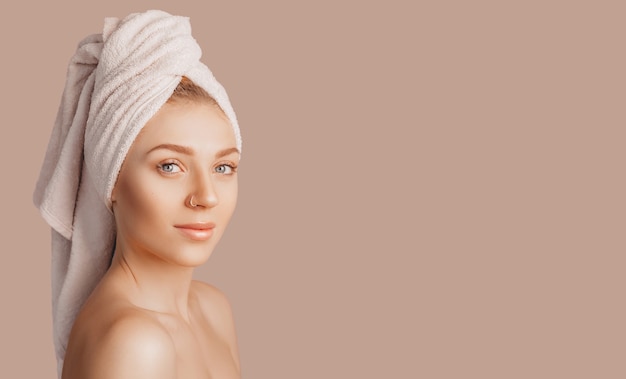 Beautiful sensual young girl with clean skin on a beige background with a mockup. Topless woman in a towel. The concept of spa treatments, natural beauty and care, youth, cream and mask, freshness