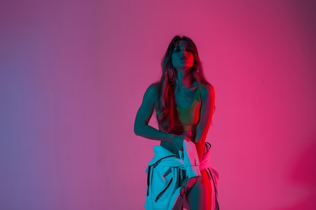 La bellissima modella sensuale della giovane donna raddrizza la felpa con cappuccio e gode di un colore incredibile al neon blu-viola in studio. la ragazza sexy in una parte superiore alla moda posa in una stanza con luce multicolore. moda da discoteca.