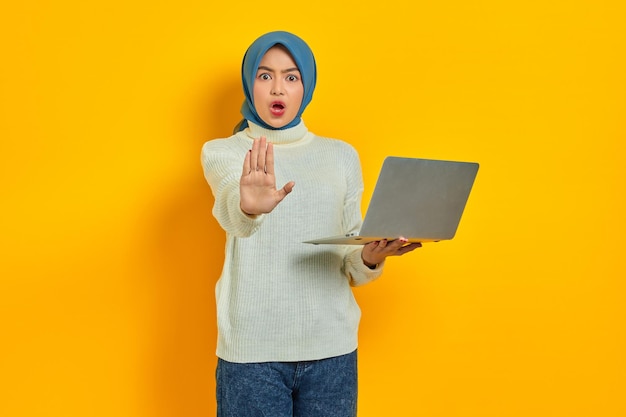 Beautiful Serious Asian woman in white sweater holding laptop with outstretched hand showing stop gesture isolated isolated over yellow background people religious lifestyle concept