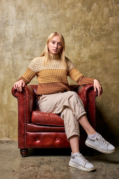 Beautiful serious bossy blonde female on leather couch isolated over brown background, in studio. posing