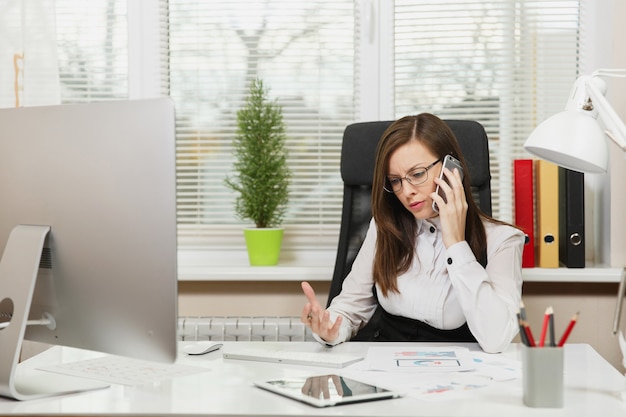 정장을 입고 안경을 쓰고 책상에 앉아 있는 아름다운 진지한 비즈니스 여성, 가벼운 사무실에서 문서를 가지고 현대적인 컴퓨터에서 일하고, 문제를 해결하는 휴대폰으로 이야기하고, 옆을 바라보고 있습니다.