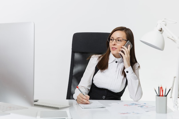 Beautiful serious business woman in suit and glasses sitting at the desk, working at contemporary computer with documents in light office, talking on mobile phone resolving issues,