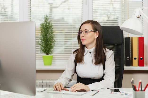 La bella donna d'affari dai capelli castani seria e assorta in giacca e occhiali seduta alla scrivania con tablet, lavorando al computer con monitor moderno con documenti in ufficio leggero, guardando da parte