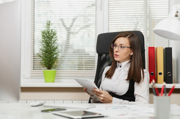 The beautiful serious and engrossed brown-hair business woman in suit and glasses sitting at the desk with tablet, working at computer with modern monitor with documents in light office, looking aside