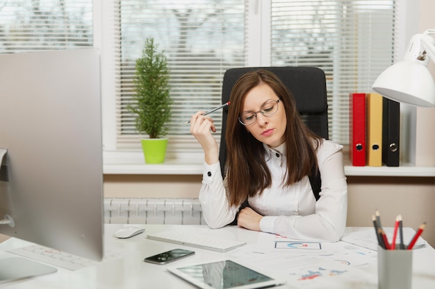 The beautiful serious and engrossed brown-hair business woman in suit and glasses sitting at the desk with tablet, working at computer with modern monitor with documents in light office, looking aside