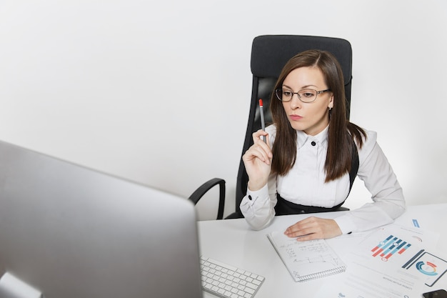 정장을 입고 안경을 쓰고 책상에 앉아 있는 아름답고 진지하고 몰두한 갈색 머리 비즈니스 여성, 밝은 사무실에서 문서가 있는 현대적인 모니터로 컴퓨터 작업,