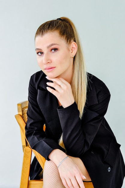 Beautiful serious girl in a black jacket sits on a chair on a gray background