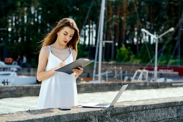 Beautiful, serious girl in a white dress with a tablet, pen, laptop phone on the dock. High quality photo