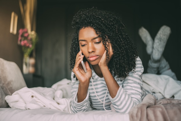 Beautiful serious thoughtful and sad black woman with curly hair using smartphone on bed